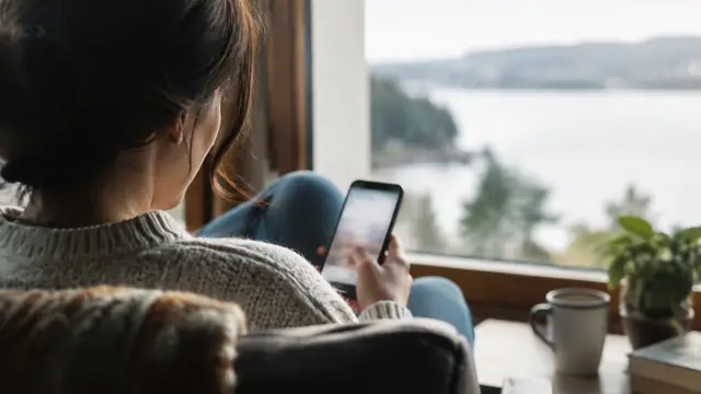 Over-the-shoulder view of a person seated by a window reviewing a smartphone, with the screen out of focus and no visible text or logos.