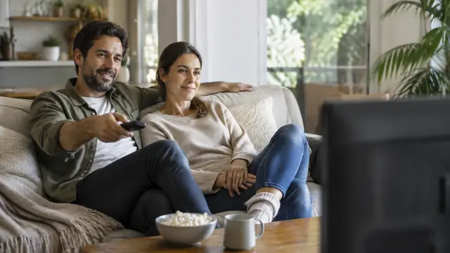 Two adults watching television together, one holding a remote, with the television facing away from the camera and no visible text, logos or brand names.