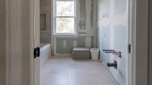 Homeowner standing in a doorway documenting an unfinished bathroom, with neatly stacked materials and capped plumbing visible.