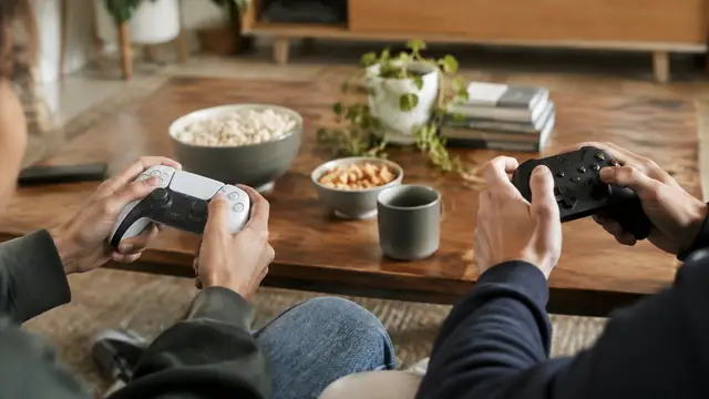 Two friends hold different styles of unbranded game controllers across a coffee table, with displays outside the frame.