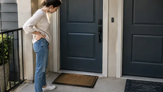 A homeowner examines an empty front doorstep beside a neighboring apartment door, with no visible house numbers, text, logos, brand names or readable screens.