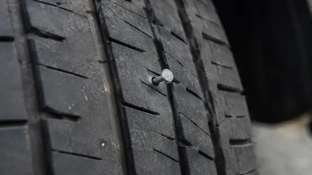 Close-up editorial photograph of a small nail lodged in the central tread of a stationary tire, with sidewall markings outside the frame and no visible branding.