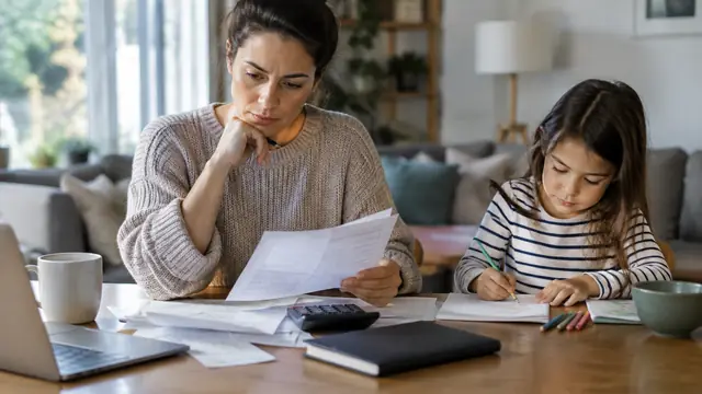 A parent reviews household finances beside a child who is quietly drawing, with a calculator and closed notebook on the table.