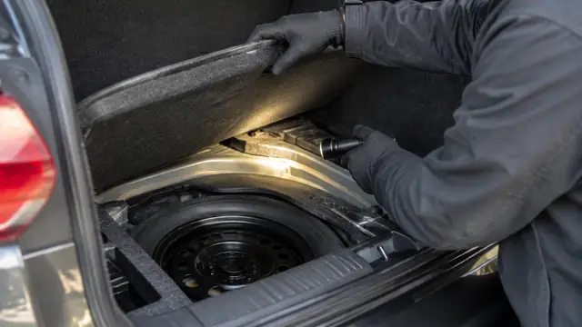 Gloved hands lift a car's trunk liner while a flashlight illuminates the spare-wheel well during an inspection for possible flood damage.