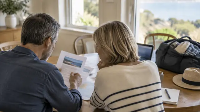 Middle-aged couple seen from behind reviewing papers at a dining table beside a packed travel bag.